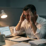 Homme stressé au bureau tard le soir, les mains sur les tempes, entouré de papiers et d'un ordinateur portable, illustrant le stress chronique lié à une élévation des enzymes hépatiques comme la GGT
